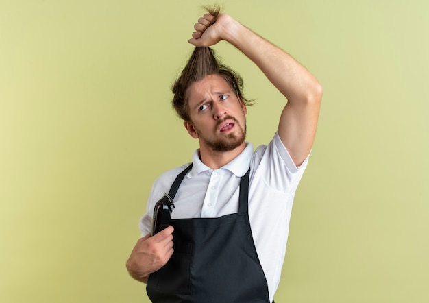 Verwirrter junger hübscher Friseur, der zur Seite schaut, die sein eigenes Haar zieht, das Haarschneidemaschinen isoliert auf olivgrün mit Kopienraum hält