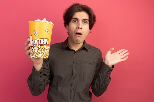Verwirrter junger hübscher Kerl, der schwarzes T-Shirt hält, das Eimer Popcorn spreizende Hand lokalisiert auf rosa Wand hält