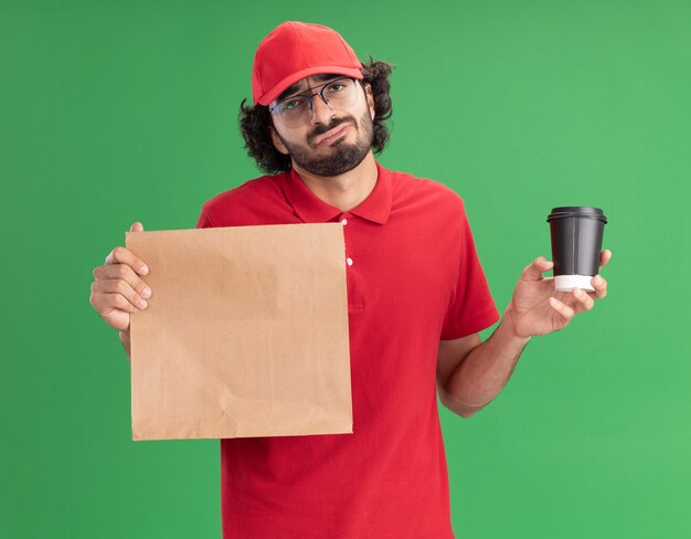 Verwirrter junger kaukasischer Liefermann in roter Uniform und Mütze mit Brille, die Papierpaket und Plastikkaffeetasse isoliert auf grüner Wand hält