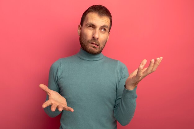 Verwirrter junger kaukasischer Mann, der nach oben schaut und leere Hände mit einem geschlossenen Auge lokal auf purpurroter Wand zeigt