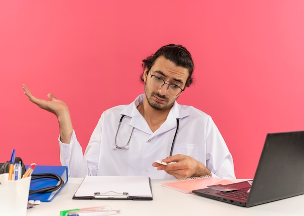 Verwirrter junger männlicher Arzt mit medizinischer Brille, der ein medizinisches Gewand mit Stethoskop trägt, das am Schreibtisch sitzt