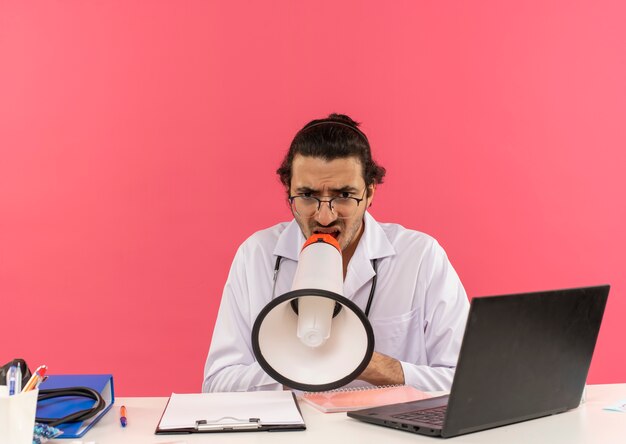 Verwirrter junger männlicher Arzt mit medizinischer Brille, der ein medizinisches Gewand mit Stethoskop trägt, das am Schreibtisch sitzt