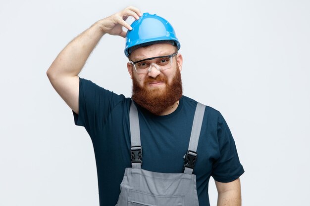 Verwirrter junger männlicher Bauarbeiter mit Schutzhelm, Uniform und Schutzbrille, der in die Kamera blickt und die Hand auf dem Helm isoliert auf weißem Hintergrund hält