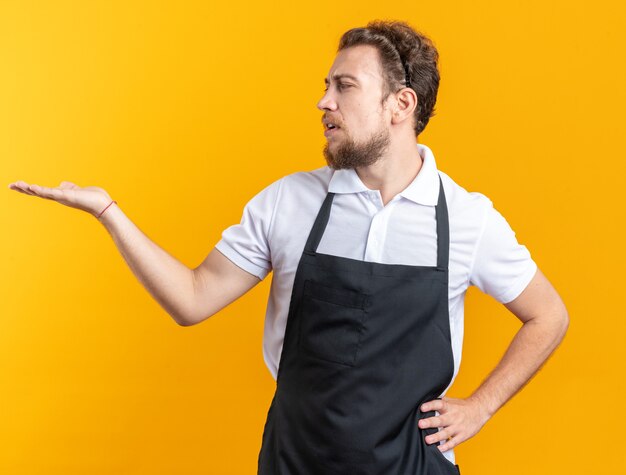 Verwirrter junger männlicher Friseur in Uniform, der vorgibt, etwas zu halten und zu sehen, das die Hand auf die Hüfte legt, isoliert auf gelber Wand