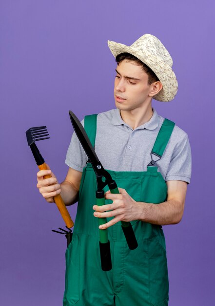Verwirrter junger männlicher gärtner, der gartenhut trägt, hält rechen und haarschneidemaschine