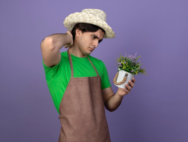 Verwirrter junger männlicher Gärtner in der Uniform, die Gartenhut hält und Blume im Blumentopf betrachtet und Hand auf Hals lokalisiert auf Purpur betrachtet