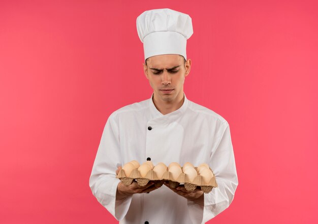 Verwirrter junger männlicher Koch, der Kochuniform trägt, die Charge von Eiern in seiner Hand auf isolierter rosa Wand betrachtet