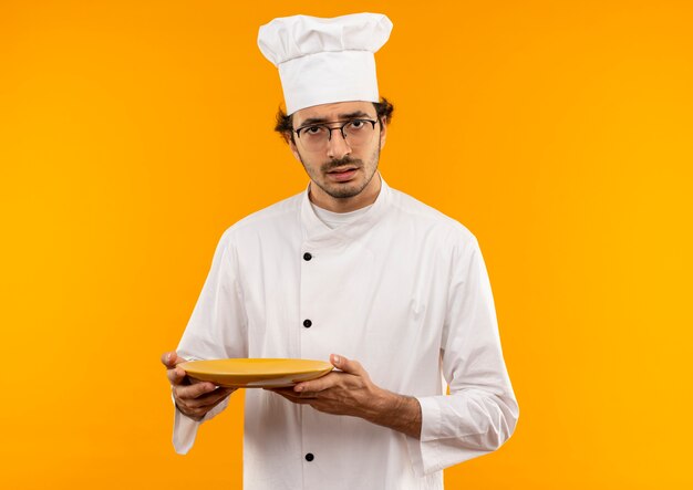 Verwirrter junger männlicher Koch, der Kochuniform und Brillenteller trägt