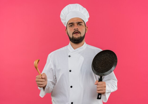 Verwirrter junger männlicher Koch in Kochuniform mit Bratpfanne und Löffel isoliert auf rosa Wand