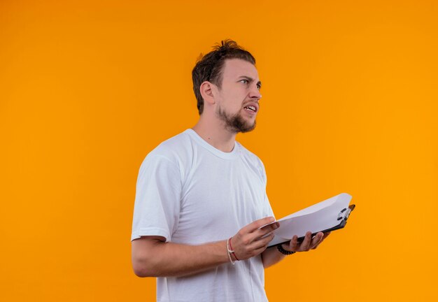 Verwirrter junger Mann, der weißes T-Shirt hält, das Klemmbrett auf isolierter orange Wand hält