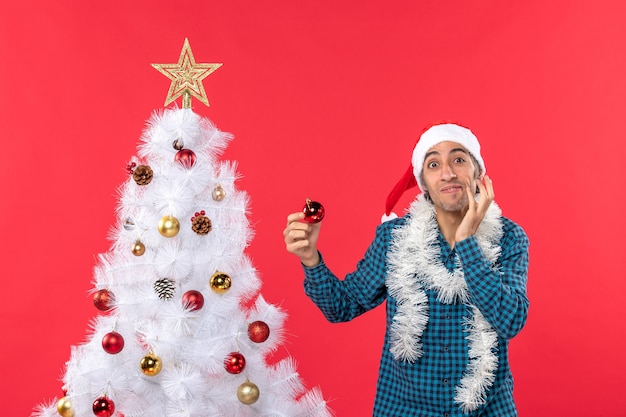 Verwirrter junger Mann mit Weihnachtsmannhut in einem blau gestreiften Hemd und Dekorationszubehör