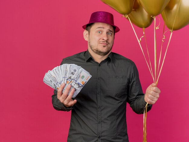 Verwirrter junger Party-Typ, der rosa Hut hält, der Bargeld mit Luftballons hält, die auf rosa lokalisiert werden