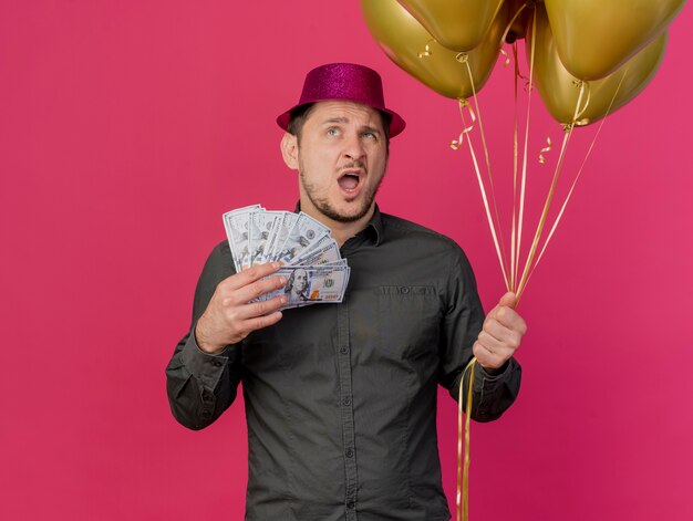Verwirrter junger Party-Typ, der rosa Hut hält, der Luftballons mit Bargeld auf Rosa isoliert hält