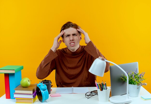 verwirrter junger Student, der am Schreibtisch mit Schulwerkzeugen sitzt, die Hände auf die Stirn legen