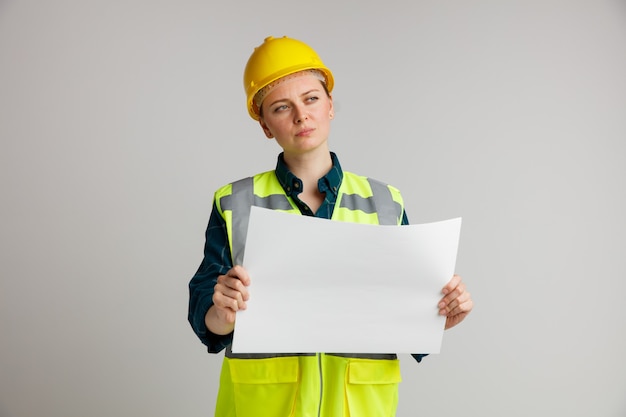 Verwirrter junger weiblicher Bauarbeiter, der Sicherheitshelm und Sicherheitsweste hält, die Papier hält, das Seite betrachtet