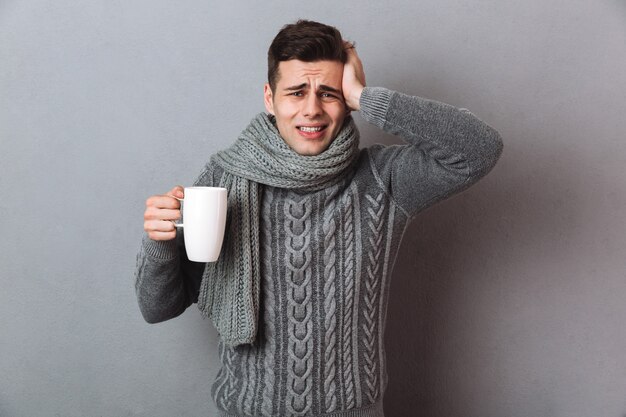 Verwirrter kranker Mann in der Strickjacke und in Schal, die Kopfschmerzen beim Halten der Tasse Tee und Schauen haben