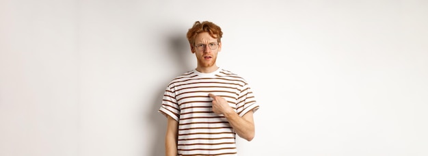 Kostenloses Foto verwirrter rothaariger mann mit brille, der auf sich selbst zeigt und den weißen hintergrund der kamera befragt