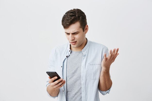 Verwirrter schockierter emotionaler junger kaukasischer männlicher Student, der verwirrten Ausdruck hatte, nachdem er Nachricht empfangen hatte, auf den Bildschirm seines Smartphones schaute, Wi-Fi benutzte und sein Gesicht in Missverständnissen runzelte.