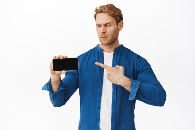 Verwirrter und zweifelnder junger Mann, der misstrauisch auf den Smartphone-Bildschirm zeigt, der etwas Seltsames auf dem Handy zeigt, das verwirrt vor weißem Hintergrund steht