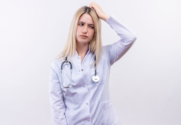 Verwirrtes Doktor junges blondes Mädchen, das Stethoskop und medizinisches Kleid trägt, legte ihre Hand auf Kopf auf isolierte weiße Wand