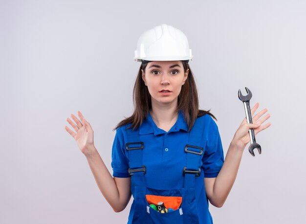 Verwirrtes junges Baumeistermädchen mit weißem Schutzhelm und blauer Uniform hält Werkstattschlüssel auf lokalisiertem weißem Hintergrund mit Kopienraum