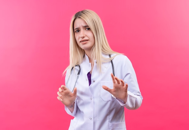 Verwirrtes junges blondes Mädchen des Arztes, das Stethoskop im medizinischen Kleid trägt, hob Hände auf isolierter rosa Wand