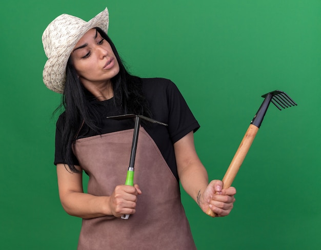 Verwirrtes junges Gärtnermädchen in Uniform und Hut mit Rechen und Hacke, das auf der grünen Wand isoliert auf den Rechen schaut