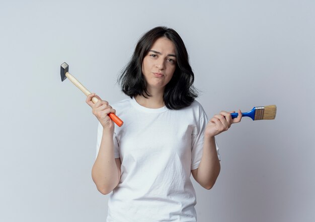 Verwirrtes junges hübsches kaukasisches Mädchen, das Hammer und Pinsel lokalisiert auf weißem Hintergrund hält