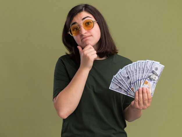 Verwirrtes junges hübsches kaukasisches Mädchen in der Sonnenbrille legt Hand auf Kinn und hält Geld auf Olivgrün