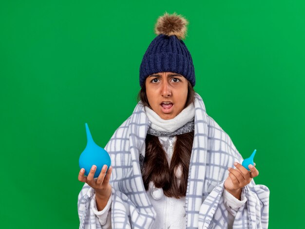 Verwirrtes junges krankes Mädchen, das Wintermütze mit Schal hält Einläufe lokalisiert auf grünem Hintergrund trägt
