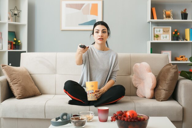 Verwirrtes junges Mädchen mit Popcorn-Eimer, das TV-Fernbedienung hält, sitzt auf dem Sofa hinter dem Couchtisch im Wohnzimmer