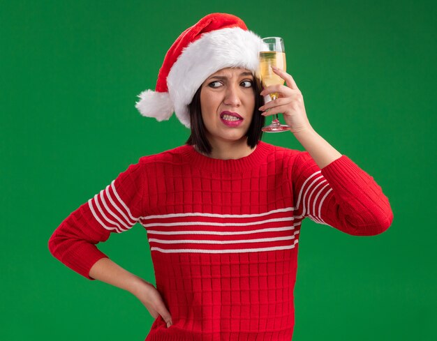 Kostenloses Foto verwirrtes junges mädchen mit weihnachtsmütze, das ein glas champagner hält und den kopf berührt, wobei die hand auf der taille gehalten wird und die seite isoliert auf grünem hintergrund betrachtet