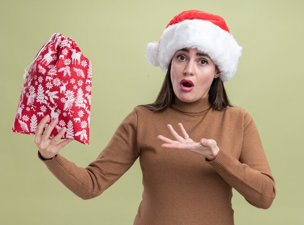 Verwirrtes junges schönes Mädchen, das Weihnachtshut hält und Punkte mit Hand an der Weihnachtstasche lokalisiert auf olivgrünem Hintergrund trägt