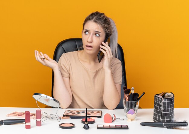 Verwirrtes junges schönes Mädchen sitzt am Tisch mit Make-up-Tools, die Make-up-Pinsel halten, spricht am Telefon, das die Hand isoliert auf oranger Wand ausbreitet