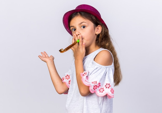 verwirrtes kleines kaukasisches Mädchen mit lila Partyhut, der Partypfeife bläst und die Hand isoliert auf weißer Wand mit Kopienraum offen hält