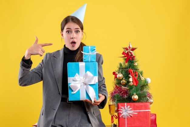 Verwirrtes Mädchen der Vorderansicht mit Partykappe, die Weihnachtsgeschenke nahe Weihnachtsbaum und Geschenkcocktail hält