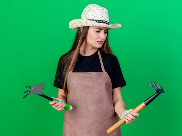 Verwirrtes schönes Gärtnermädchen in der Uniform, die Gartenhut trägt, der Hacke Rechen hält und Rechen in ihrer Hand lokalisiert auf grünem Hintergrund betrachtet