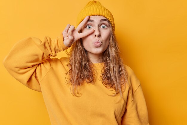 Verwundertes tausendjähriges Mädchen mit langen Haaren hält die Lippen gefaltet, macht eine Friedensgeste über dem Auge, fühlt sich beeindruckt, gekleidet in Freizeitkleidung, isoliert auf leuchtend gelbem Hintergrund, genießt das Konzept der Körpersprache