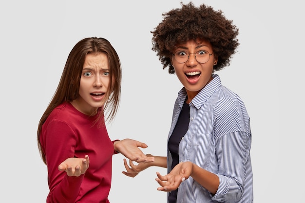 Kostenloses Foto verzweifelt frustriert breiteten zwei gemischte frauen die hände aus, standen seitwärts, schauten empört, modell gegen weiße wand. also was zu tun? verwirrte multiethnische mädchen schreien laut