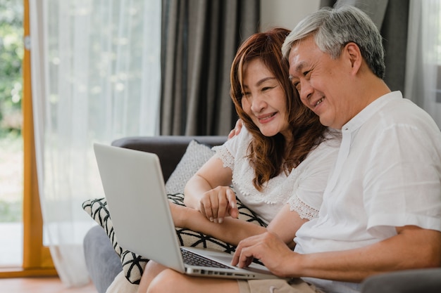 Videoanruf der asiatischen älteren Paare zu Hause. Asiatische ältere chinesische Großeltern, unter Verwendung des Laptopvideoanrufs, der mit Familienenkelkind spricht, scherzt beim auf Sofa im Konzept des Wohnzimmers zu Hause liegen.