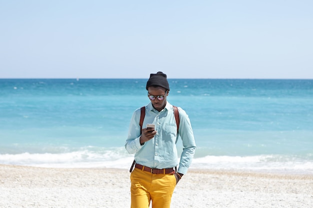 Vielbeschäftigter junger dunkelhäutiger Europäer, der modische, trendige Kleidung und einen Rucksack trägt und auch in den Ferien online bleibt, am Strand ein Mobiltelefon benutzt und alle Schönheiten ignoriert, die ihn umgeben