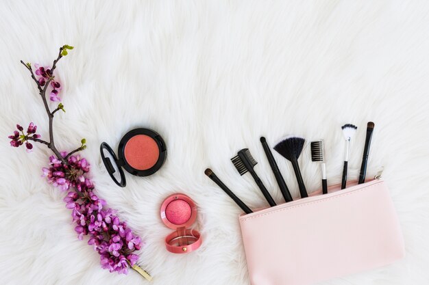 Viele Make-up-Pinsel aus rosa Tasche; Blumenzweig und kompaktes Gesichtspuder auf weichem weißem Fell