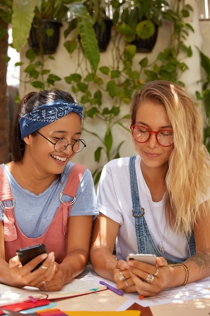 Viele multiethnische Frauen teilen Multimediadateien über Bluetooth, halten moderne Mobiltelefone, sitzen am Desktop, arbeiten für gemeinsame Aufgaben zusammen, tragen transparente Brillen und sind süchtig nach modernen Technologien