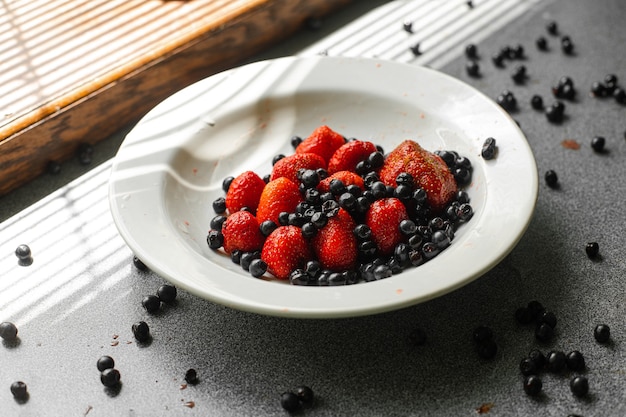 Viele saftige frische reife rote Erdbeeren und Brombeeren liegen in einer weißen Keramikplatte auf dem Tisch unter hellem Sonnenlicht