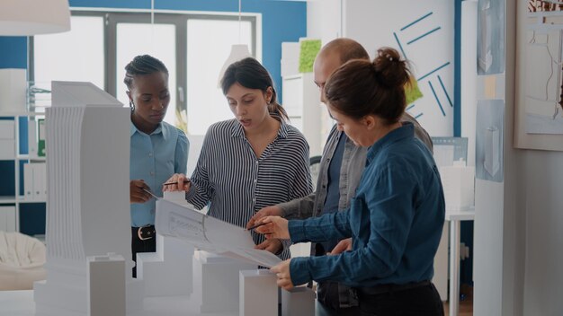 Vielfältige Gruppe von Kollegen, die an der Konstruktion von Modellen mit Bauplänen arbeiten. Mitarbeiter, die maquette und industrielle skizzen verwenden, um architekturprojekte zu erstellen und ideen zu brainstormen.