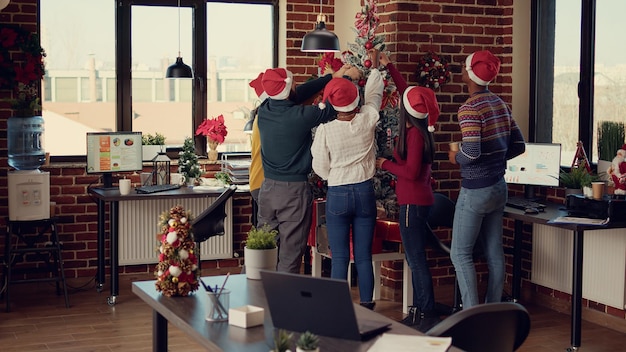 Vielfältige Gruppe von Kollegen schmücken den Weihnachtsbaum im Startup-Büro, fühlen sich glücklich und festlich mit Ornamenten der Wintersaison. Verwenden von Lichtern und Dekorationen, um Heiligabend zu feiern.