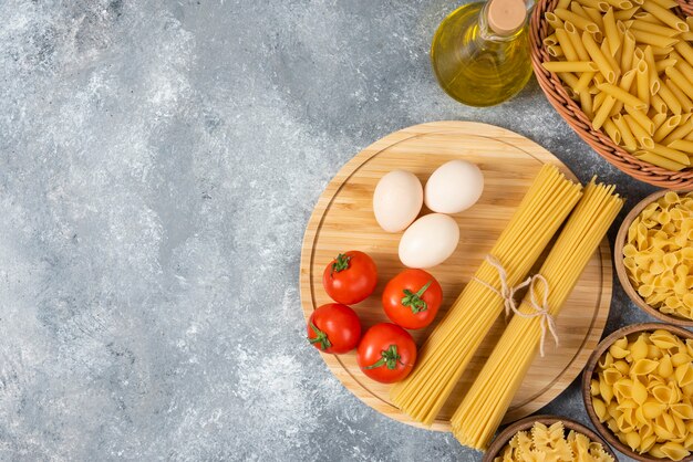Vielzahl von rohen Nudeln mit Eiern, frischen Tomaten und einer Flasche Öl auf Marmoroberfläche.