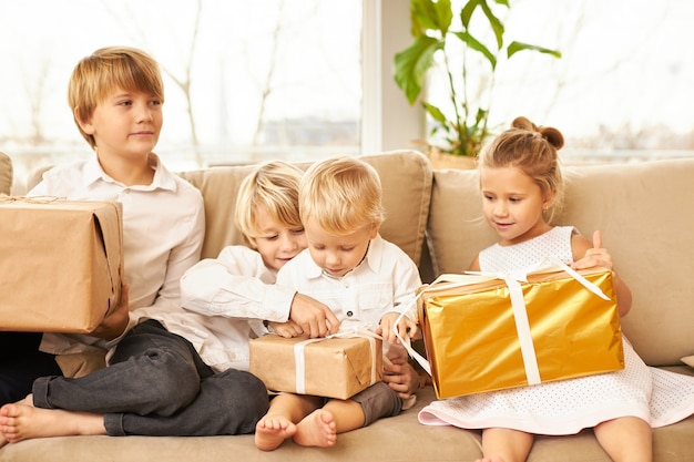 Vier kaukasische Kinder, die identische weiße Hemden und keine Socken tragen, sitzen auf dem Sofa im Wohnzimmer, sind ungeduldig, Kisten mit Neujahrsgeschenken zu öffnen, lächeln und haben freudig aufgeregte Gesichtsausdrücke