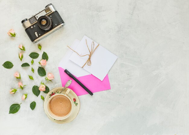 Vintage Kamera; pinke Rose; grüne Blätter; Briefumschlag; Papier; Stift und Kaffeetasse auf konkreten Hintergrund