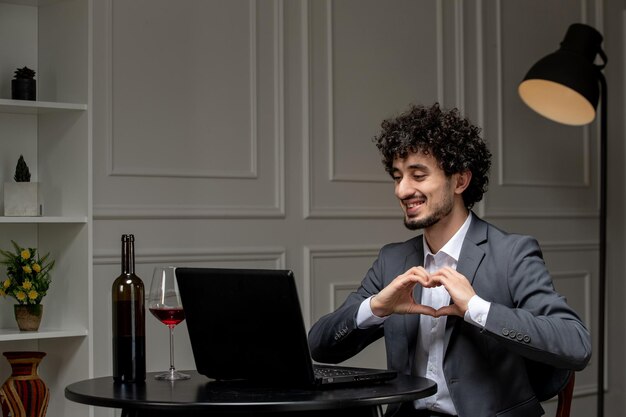 Virtuelle Liebe hübscher süßer Kerl im Anzug mit Wein auf einem Ferncomputerdatum mit Herzgeste